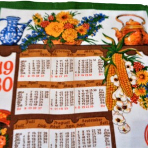 Op de afbeelding: Een witte theedoek met een bedrukte kalender van 1980. De kalender heeft maanden in het Duits en Engels. De theedoek heeft een groene rand en een bloemenmotief met een theepot en een kan.