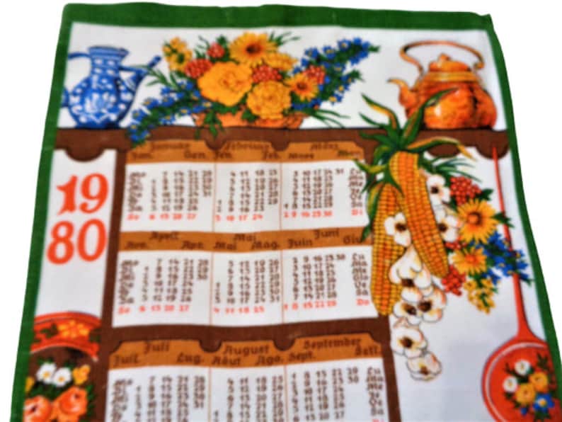 Op de afbeelding: Een witte theedoek met een bedrukte kalender van 1980. De kalender heeft maanden in het Duits en Engels. De theedoek heeft een groene rand en een bloemenmotief met een theepot en een kan.