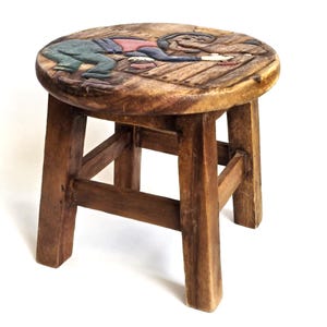 Puede incluir: Un pequeño taburete de madera con asiento redondo y cuatro patas. El asiento presenta un relieve tallado de una figura y una rana en tonos azules, rojos y marrones. El taburete es de madera oscura teñida.