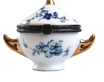 May include: A white porcelain trinket box with a blue and white floral design. The box has a lid with a gold trim and two gold handles.