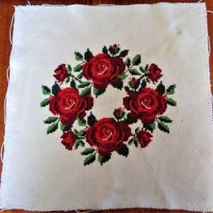 May include: White fabric with a red rose wreath embroidered in the center. The roses are surrounded by green leaves.
