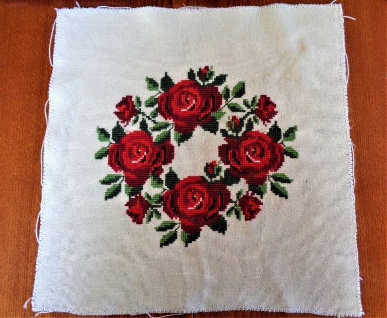 May include: White fabric with a red rose wreath embroidered in the center. The roses are surrounded by green leaves.
