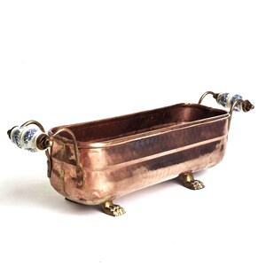 Peut inclure: Une jardinière rectangulaire en cuivre avec une finition martelée, dotée de deux poignées métalliques incurvées avec des accents en céramique florale bleue et blanche. Le récipient repose sur quatre petits pieds en laiton en forme de pattes.