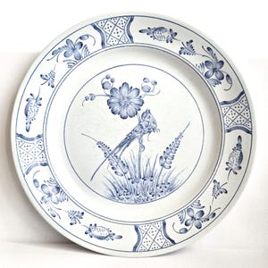 May include: White ceramic plate with a blue floral and bird design. The plate features a detailed illustration of a bird perched on a branch, surrounded by flowers and foliage. The rim has a repeating pattern of flowers and geometric shapes.