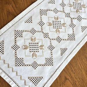 May include: White linen table runner with a delicate embroidered design in a beige color. The pattern features intricate geometric shapes and a central floral motif.