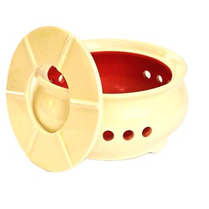 Op de afbeelding: Een witte keramische pot met een rode binnenkant en een bijpassend deksel. De pot heeft gaten aan de onderkant en zijkanten, en het deksel heeft een gat in het midden. De pot is ontworpen om voedsel te koken of te serveren.