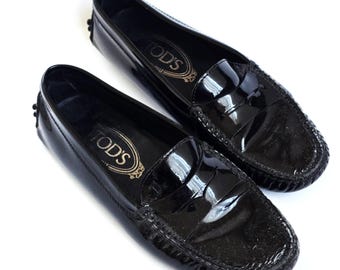 Könnte beinhalten: Ein Paar schwarze Loafer aus Lackleder mit glänzender Oberfläche. Jeder Schuh verfügt über einen klassischen Penny-Loafer-Riemen über dem Vorderblatt und das in Gold gedruckte Tod's-Markenlogo auf der Innensohle. Die Sohle hat ein Noppenprofil.