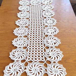 以下が含まれることがあります： 繊細な花柄の白いクロシェのテーブルランナー。ランナーは細い糸でできており、繊細でレースのような外観です。