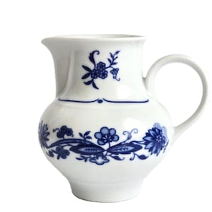 May include: White ceramic jug with a rounded body and a handle. The jug is decorated with blue floral patterns and a blue decorative line. The jug has a spout for pouring.