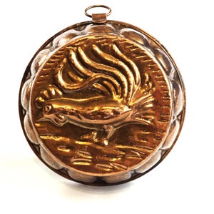 Puede incluir: Un molde de metal de color cobre vintage con un diseño de gallo. El molde redondo presenta un relieve detallado de un gallo con una cola grande y un lazo para colgar en la parte superior. El molde tiene un borde decorativo y texturizado.