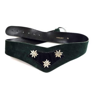May include: A dark green suede belt featuring a V-shaped front panel with three silver-toned edelweiss flower ornaments. The interior is lined with black leather, stamped with the text ECHTES LEDER and the number 90, and includes a silver buckle.