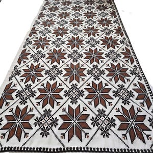 May include: A white textile with a repeating pattern of brown star-like shapes and black geometric accents. The edges are finished with a black, scalloped border. This decorative textile could be used as a table runner or wall hanging.