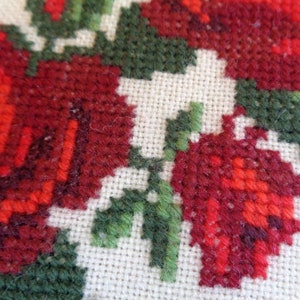 May include: Close-up of a red and green cross-stitch embroidery featuring a floral design with roses and leaves.