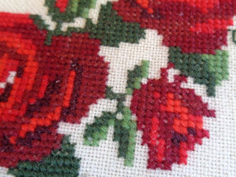 May include: Close-up of a red and green cross-stitch embroidery featuring a floral design with roses and leaves.