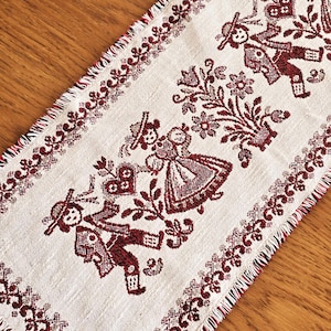 Può includere: Runner da tavolo color crema con un motivo ripetuto di figure folcloristiche e motivi floreali bordeaux. I bordi sono sfrangiati e il design presenta persone e fiori stilizzati.