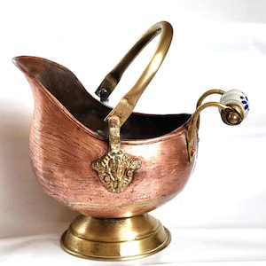 Könnte beinhalten: Ein kupferfarbener Kohleeimer mit Messinggriff und -sockel. Der gebogene Griff ist mit einer dekorativen Messinghalterung am Eimer befestigt. Der kleine Seitenbügel hat einen weißen und blauen Keramikknauf. Der Eimer hat einen Ausguss.