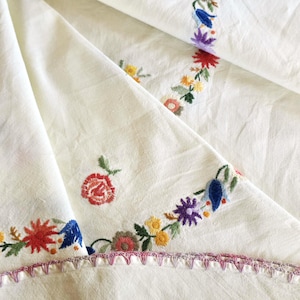 Può includere: Una tovaglia in tessuto bianco sporco caratterizzata da colorati ricami floreali nelle tonalità del rosso, blu, giallo, viola e verde. I bordi sono rifiniti con una decorazione cucita in rosa e viola, che mostra dettagli di ricamo.