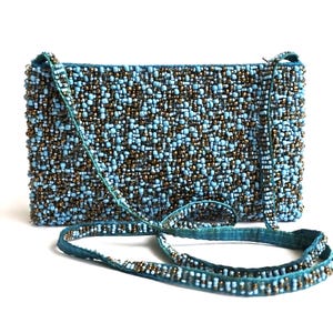 Può includere: Una borsa rettangolare con perline e una lunga tracolla. La borsa è ricoperta di piccole perline azzurre e bronzo. La tracolla è di un colore verde acqua abbinato, anch'essa ornata di perline. La borsa è posta su uno sfondo bianco.