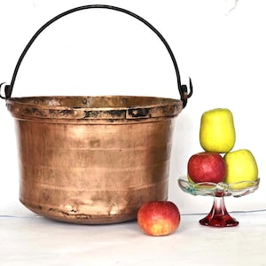 Puede incluir: Una olla grande de cobre con un asa de hierro negro. La olla está sobre una superficie blanca. Hay tres manzanas en un pedestal de vidrio al lado de la olla.