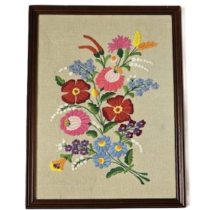 May include: A framed embroidery featuring a colorful floral bouquet with red, pink, blue, and yellow flowers. The flowers are embroidered on a beige linen fabric and framed in a dark brown wood frame.