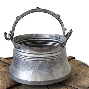 May include: A silver-toned metal bucket with a curved handle and decorative details. The bucket features horizontal bands around the body. The handle is attached with ornate, curved brackets. The bucket rests on a wooden surface.