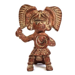 May include: A terracotta-colored figurine of a warrior with a large headdress and shield. The figure is in a defensive stance, holding a weapon. The shield has a circular design. The statue is likely a representation of ancient art.