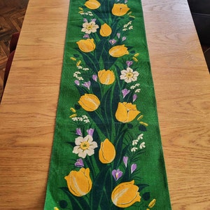 May include: A green table runner with a floral pattern featuring yellow tulips, white flowers, and purple flowers.