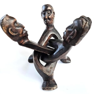 Puede incluir: Una escultura de madera oscura de tres figuras entrelazadas con rostros humanos. Las figuras están talladas en un estilo estilizado y abstracto.