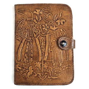 May include: A brown leather wallet with a detailed embossed design. The design features figures in ornate clothing and floral elements. The wallet has a snap closure and is rectangular in shape.