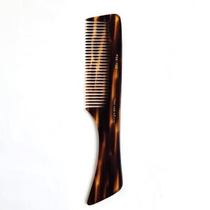 Pode incluir: Um pente de cabelo com padrão de tartaruga em tons de castanho e preto. O pente apresenta dentes finos na parte superior e um cabo sólido. O texto dourado impresso na lateral diz PAX 150 e HAND MADE IN SWEDEN. O design é liso e uniforme.
