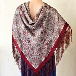 May include: A shawl with a paisley pattern in shades of red, gray, and beige. The shawl has a deep red border and fringe. The shawl is draped over the shoulders.
