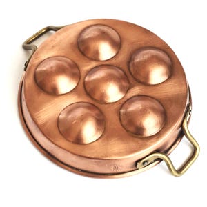 May include: Copper escargot pan with six round indentations. The pan is circular with a raised rim and two brass handles. The copper has a warm, reddish-brown colour, and the handles are a contrasting gold.