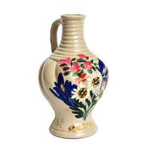 Puede incluir: Una jarra de cerámica blanca con un diseño floral pintado en azul, rosa, verde y amarillo. La jarra tiene un asa y un cuerpo redondo. La parte inferior de la jarra tiene el texto "G. und F. 1901" y "Ger. aus Brugg."