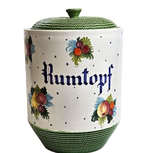 May include: A white ceramic Rumtopf jar with a green lid and base. The jar is decorated with colourful fruit illustrations and the word "Rumtopf" in blue script. The lid has a textured pattern, and the jar has a rounded shape.