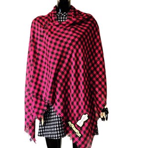 May include: A lightweight scarf featuring a vibrant hot pink and black buffalo check pattern. The fabric has a soft, draped appearance with frayed edges along the bottom hem. A small white patch with a black outline is attached to the lower right corner.