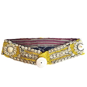 Peut inclure: Une ceinture en perles blanches et jaunes avec un grand bouton en coquille blanche. La ceinture est faite d'un tissu tissé avec un motif bleu foncé et rouge.