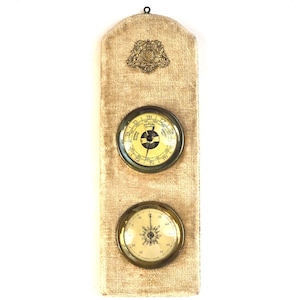May include: A vintage wall-mounted weather station with a beige fabric backing. It features two circular gauges with brass-coloured frames and a decorative emblem at the top. The gauges display temperature and barometric pressure readings.