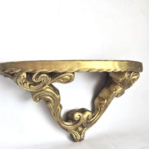 May include: A gold-colored wall shelf with ornate, swirling details. The shelf has a rounded front edge and decorative supports. The design is reminiscent of antique or vintage decor.