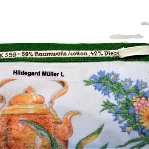 Op de afbeelding: Een witte theedoek met een groene rand en een bloemenpatroon. De theedoek heeft een theepot en een boeket blauwe, gele en roze bloemen. De tekst "WK 559 - 58% Baumwolle/cotton, 42% Viscose" staat op de rand gedrukt. De tekst "Hildegard M&uuml;ller L" staat op een wit label gedrukt.