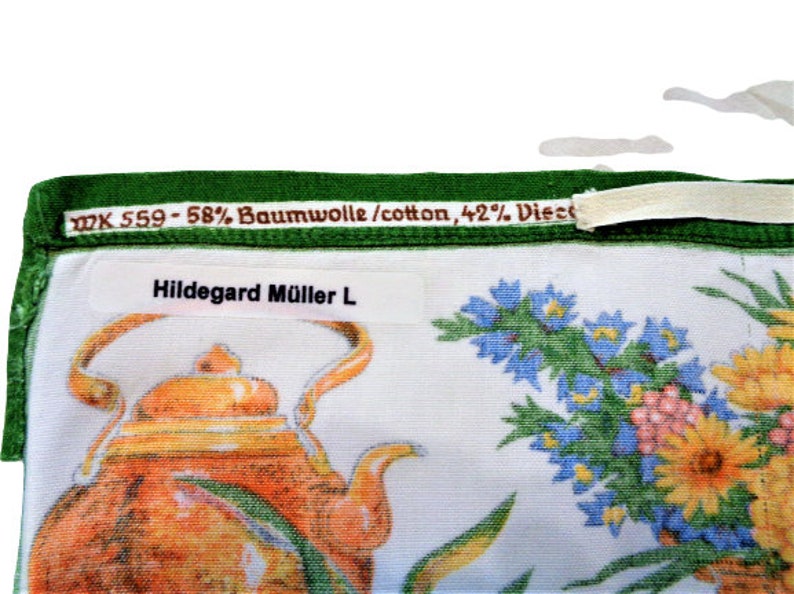 Op de afbeelding: Een witte theedoek met een groene rand en een bloemenpatroon. De theedoek heeft een theepot en een boeket blauwe, gele en roze bloemen. De tekst "WK 559 - 58% Baumwolle/cotton, 42% Viscose" staat op de rand gedrukt. De tekst "Hildegard M&uuml;ller L" staat op een wit label gedrukt.