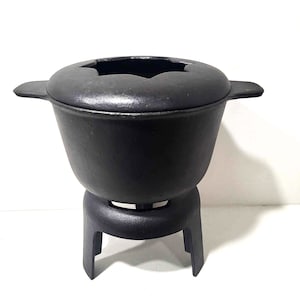 Puede incluir: Olla de fondue de hierro fundido negro con una abertura en forma de estrella en la tapa y tres patas.
