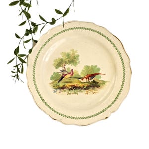 May include: A round, cream-colored ceramic plate with a scalloped edge and a green leaf-patterned border. The center features a colorful illustration of two birds in a landscape with green trees and bushes against a light background.