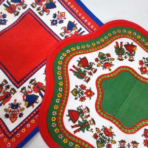 Può includere: Due tovagliette all'americana in tessuto rosso e bianco con un design di arte popolare che presenta coppie in abiti tradizionali. Le tovagliette hanno un bordo verde e bianco con un bordo smerlato.