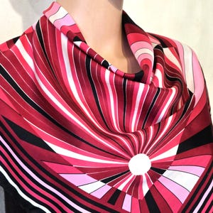 Può includere: Una sciarpa vibrante con un motivo a strisce nei toni del rosso, rosa e bianco. Il design presenta strisce radiali che emanano da un cerchio bianco centrale. La sciarpa è drappeggiata, mostrando il suo motivo geometrico e la tavolozza dei colori.