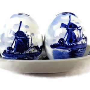 Może przedstawiać: Zestaw solniczki i pieprzniczki z ceramiki w kolorze niebieskim i białym, w kształcie jajek z motywem wiatraka. Solniczki stoją na dopasowanej owalnej podstawce.