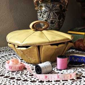 May include: A vintage, pale yellow, leatherette sewing kit with a handle, open to reveal the interior. A pink measuring tape, spools of thread, and a small container of black buttons are nearby. A decorative vase is in the background.