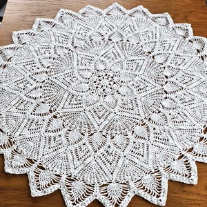 May include: A white, circular, crocheted doily with a detailed, openwork design. The doily features a central floral motif surrounded by geometric patterns and a scalloped edge. The handmade textile is displayed on a wooden surface.