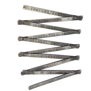 Pode incluir: Uma régua dobrável antiga de metal, com marcações de medição visíveis. A régua prateada está disposta em zigue-zague sobre um fundo branco. As medidas são em centímetros.