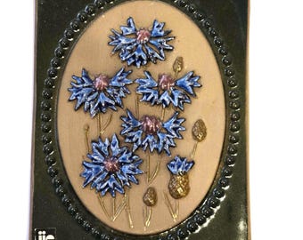 May include: A rectangular ceramic tile with a raised relief design of blue cornflowers with pink centres and gold stems. The flowers are set against a tan oval background, framed by a dark green border with a beaded edge. The tile has the text "jie" and "Gantofta Sweden".