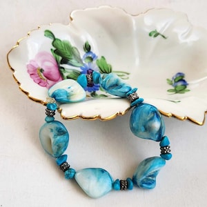 Könnte beinhalten: Ein türkisfarbenes und weißes Muschelperlenarmband mit silbernen Akzenten. Das Armband liegt auf einer weißen Porzellanschale mit Blumenmuster.